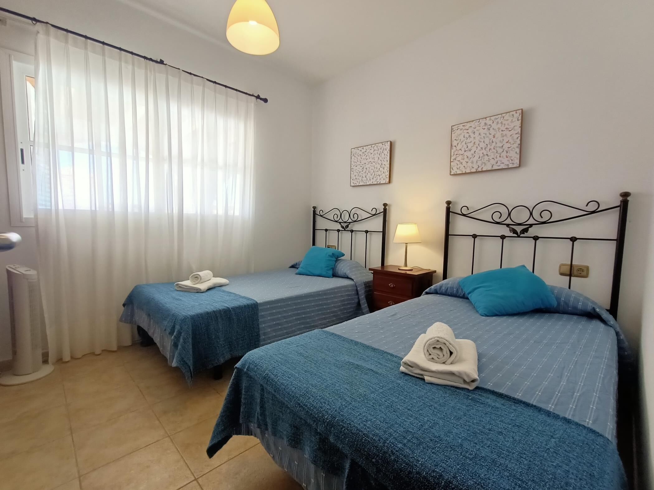Ein Zimmer mit zwei Betten, jeweils mit blauer Bettdecke und Kissen, einem Nachttisch mit Lampe sowie einem Fenster mit durchsichtigen Vorhängen.