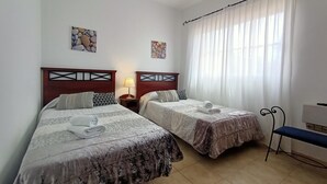 Una camera d'albergo con due letti, un comodino con lampada, una panca e una finestra con tende.
