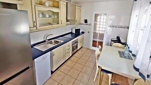A compact kitchen with a refrigerator, sink, microwave, and cabinets. There is a dining table with chairs and a window with curtains.