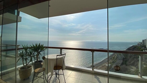 Ein Balkon mit Tisch und Stühlen, der auf eine Küstenlandschaft mit Meer und entfernten Gebäuden blickt.