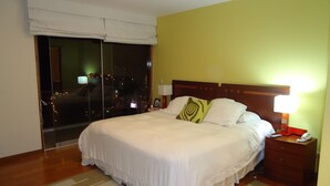 Ein Hotelzimmer mit einem großen Bett, einem Nachttisch mit Lampe, einem Fenster mit Blick auf die beleuchtete Stadt und einer Schiebetür.