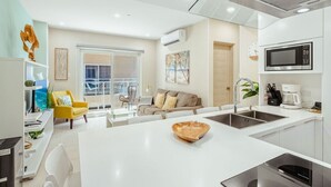 Una cocina moderna con una isla central, un comedor con una silla amarilla y una sala de estar con sofá y un ventanal grande.