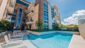 Un hotel moderno con piscina, varios balcones y un cielo azul despejado.