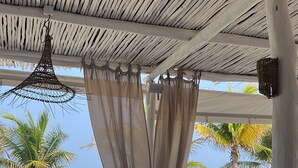 Ein Außenbereich mit weißen Tischen und Stühlen, einer gewobenen Decke und Blick auf Strand und Meer.