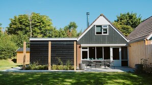 A modern house with a dark exterior, a sloped roof, and a large sliding glass door leading to a patio with outdoor furniture. - Modern Holiday Home With Spacious Garden and Terrace Suitable for 5 People (Ouddorp)