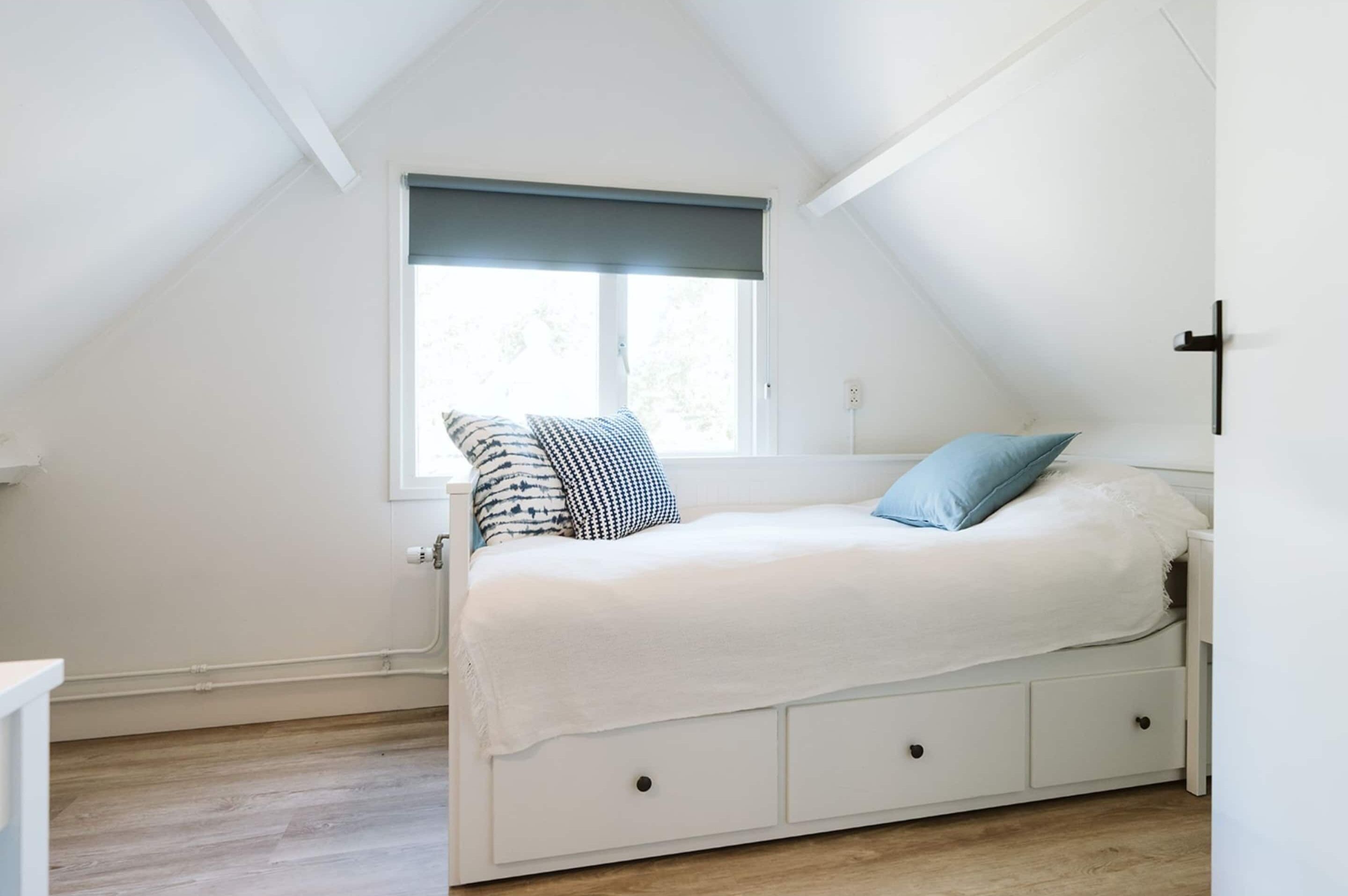 Une chambre avec un plafond en pente, une fenêtre munie d’un store foncé, un lit avec un matelas blanc et deux oreillers, et une table de chevet blanche.