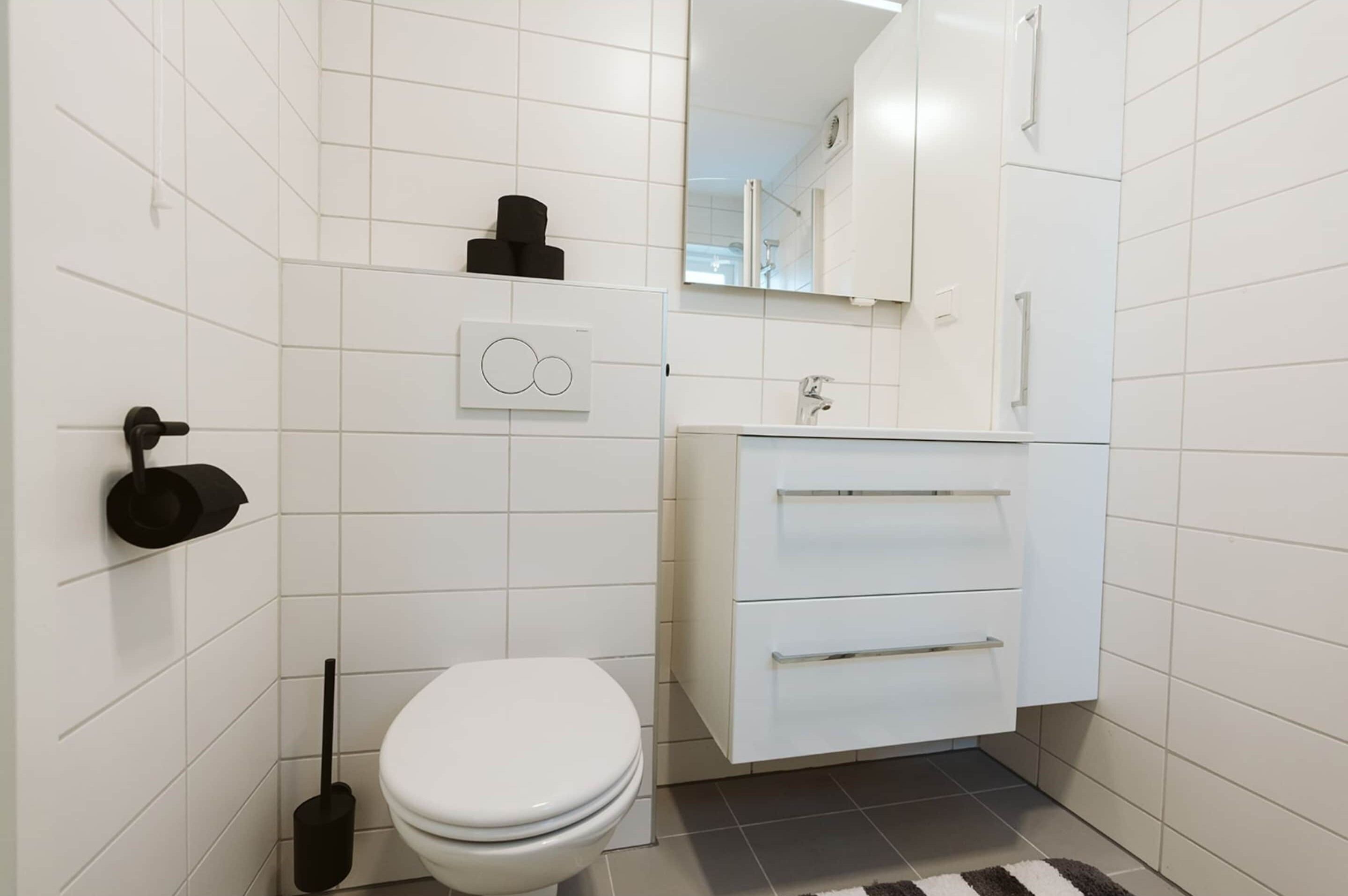 Een moderne badkamer met witte tegels, een toilet, een wastafel met spiegel en een toiletrolhouder.