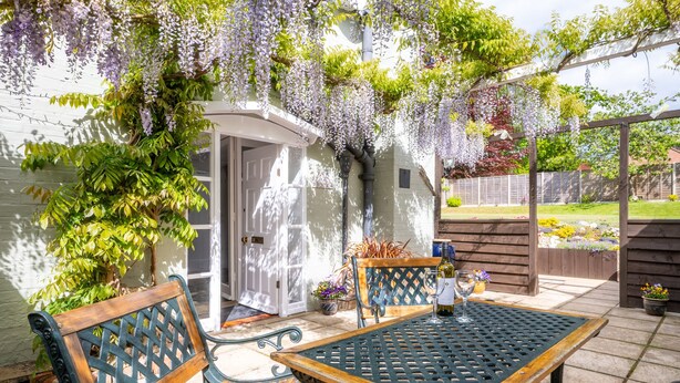 Een terras buiten met een tafel en twee stoelen, omringd door weelderig groen en een pergola met bloeiende planten.