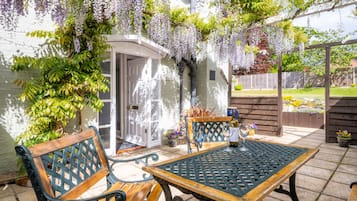 An outdoor patio with a table and two chairs, surrounded by lush greenery and a pergola with hanging flowers.
