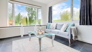 A modern living room with a glass coffee table, a grey sofa with patterned cushions, and large windows offering a view of greenery. - Water Villa in Ooltgensplaat Near Volkerak (Ooltgensplaat)