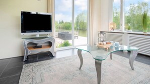 A modern living room with a flat-screen TV, a glass coffee table, and a view of greenery through large windows. - Water Villa in Ooltgensplaat Near Volkerak (Ooltgensplaat)