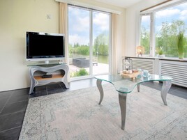 Uma sala de estar moderna com televisão de tela plana, uma mesa de centro de vidro e vista para o verde através de grandes janelas.
