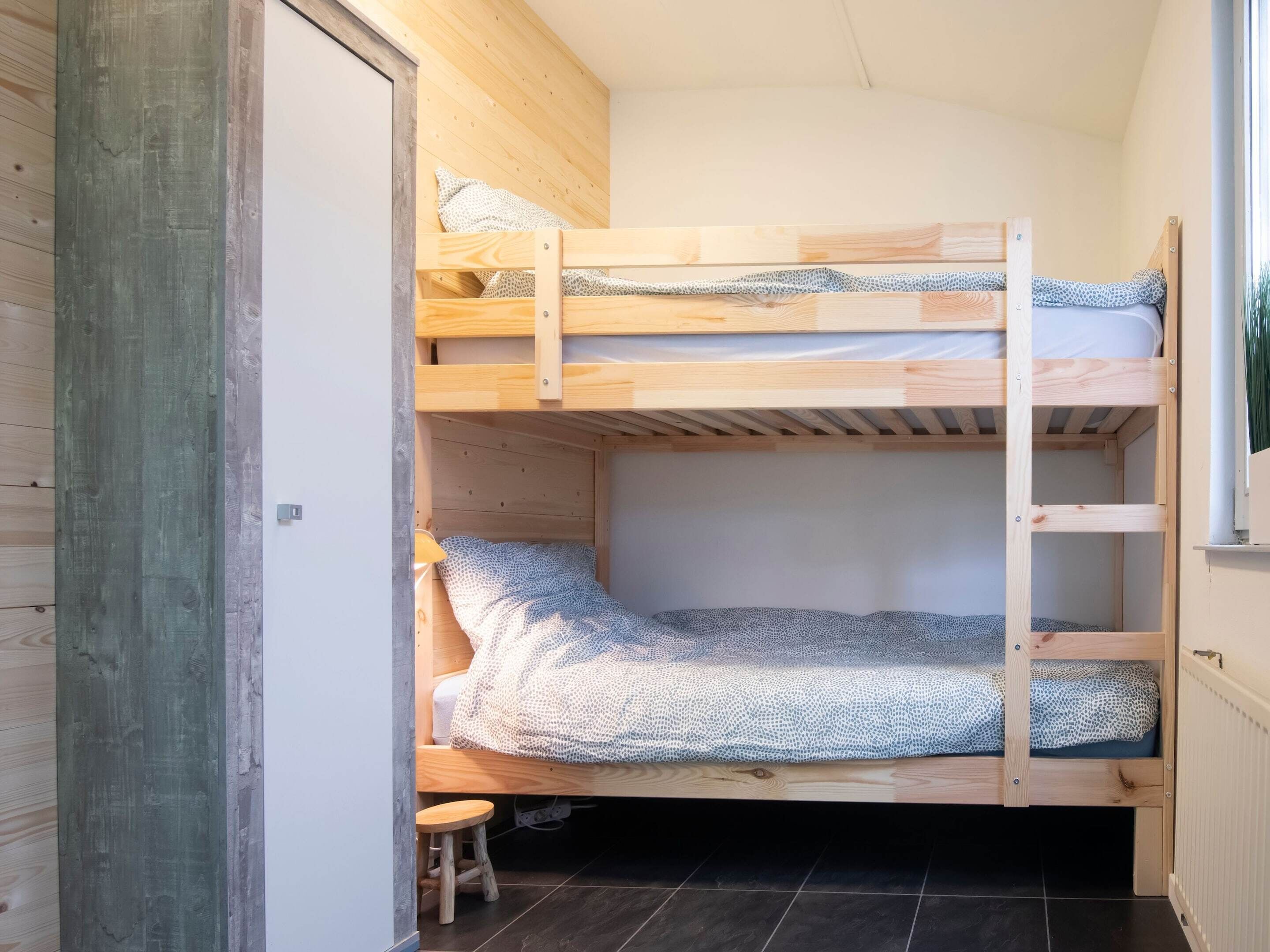 A bunk bed with a wooden frame, a small stool, and a window with a view of greenery.