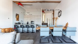 A modern kitchen with a central island, bar stools, and a dining area with a wooden table and blue chairs. - Spacious Luxury Apartment With Beautiful Views of the Harbor and the North Sea (Ouddorp)