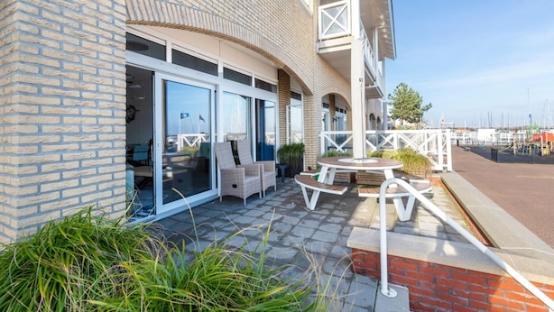 A building with a brick facade, large glass doors, outdoor seating area with tables and chairs, and a view of the marina in the background. - Spacious Luxury Apartment With Beautiful Views of the Harbor and the North Sea (Ouddorp)