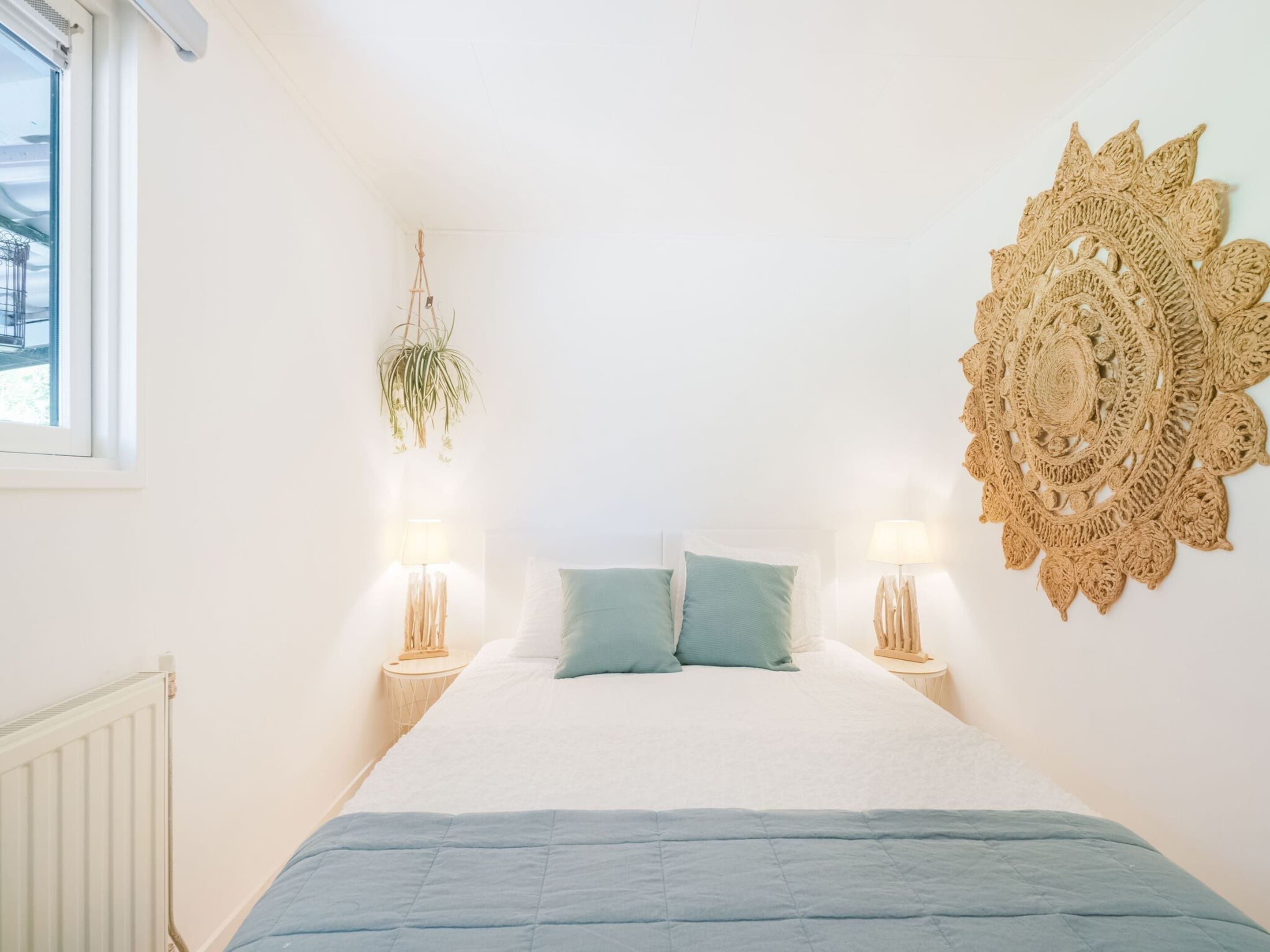 A bedroom with a bed, two bedside lamps, a hanging plant, and a decorative wall piece.