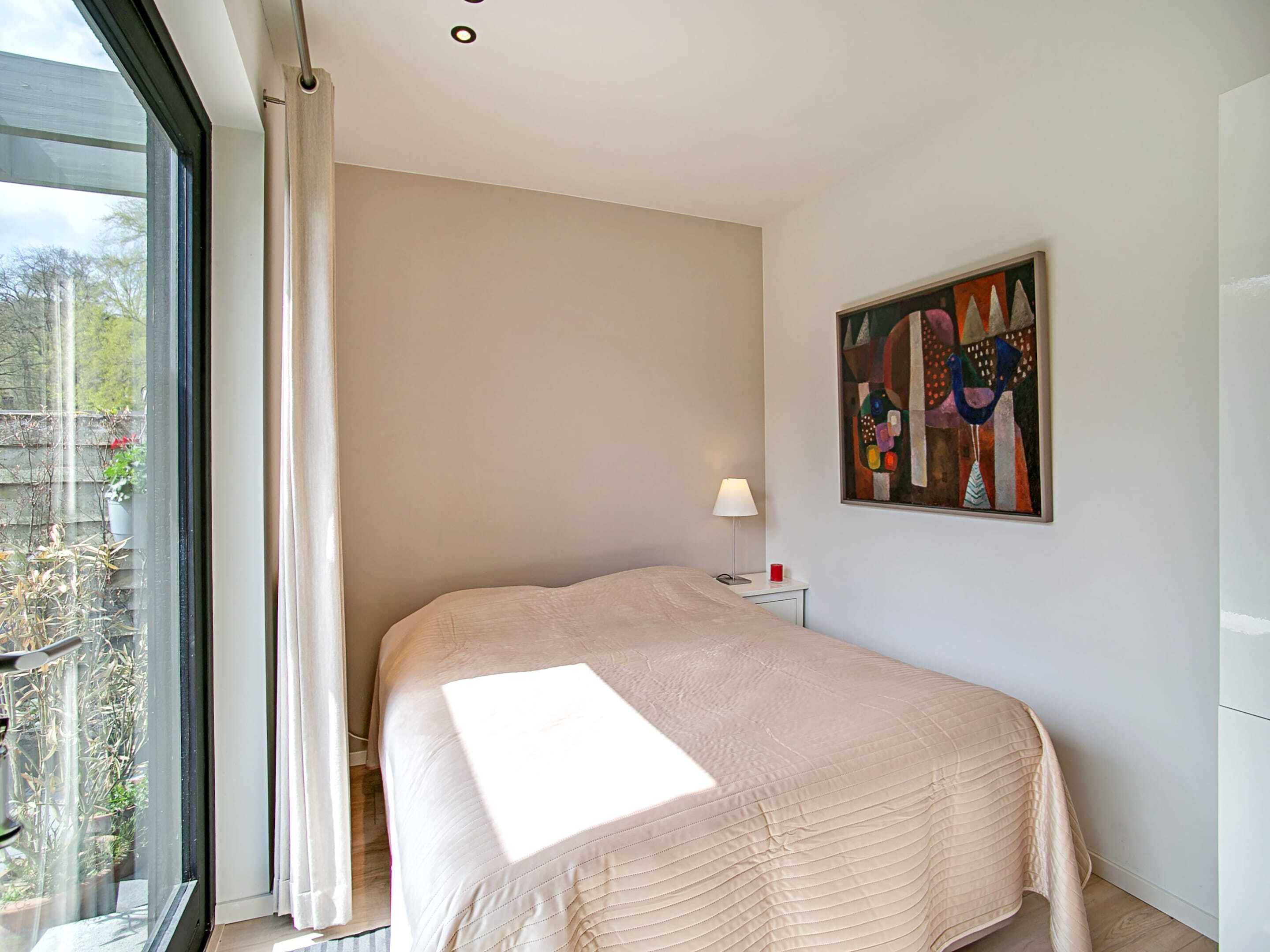 Une chambre à coucher avec un lit, une table de chevet avec une lampe, un tableau au mur et une fenêtre avec des rideaux.