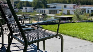 A metal bench with a slatted seat and backrest, positioned on a paved area with a grassy lawn and a building in the background. - Villa in Harderwold With Garden (Zeewolde)
