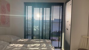A bedroom with a bed, a chair, and a view through a window with blinds. - Villa in Harderwold With Garden (Zeewolde)
