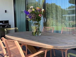 Et rundt træbord med en glasvase med blomster, omgivet af stole, i et rum med udsigt til træer gennem et vindue.