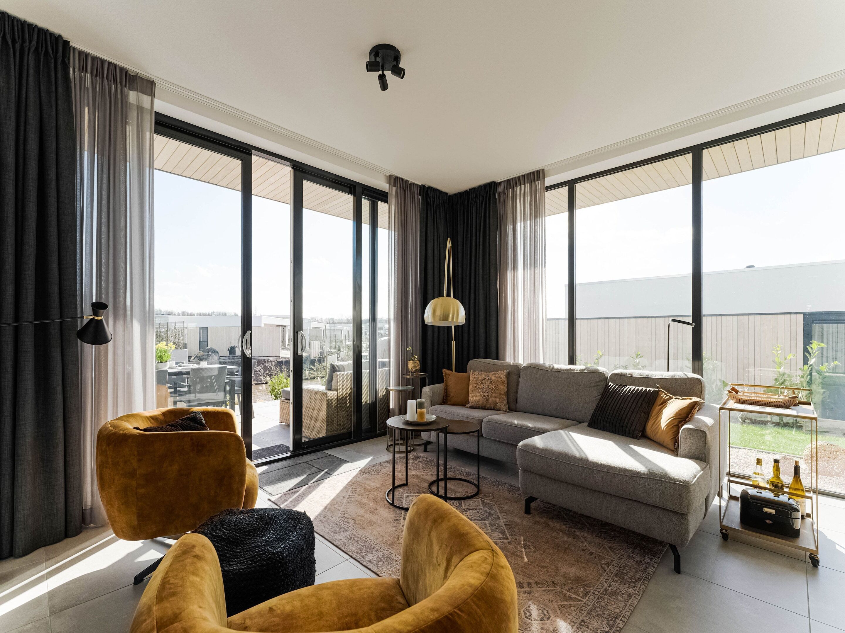 A spacious living room with a grey sofa, a yellow armchair, and a black ottoman. Large windows offer a view of the outdoors.