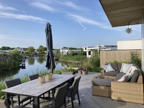 An outdoor patio with a table and chairs, a covered seating area, and a view of a lake and buildings in the background. - Villa in Zeewolde With Private Jetty (Zeewolde)