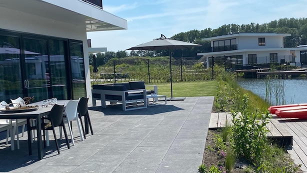 Un patio moderno al aire libre con zona de comedor, piscina y vista a un lago y edificios al fondo.