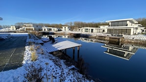 Una zona residencial moderna con una gran masa de agua que refleja los edificios y el cielo. Varias casas modernas con ventanas amplias y un muelle que se adentra en el agua.