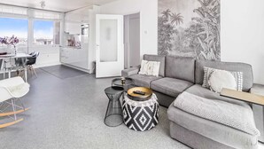 A modern living room with a grey sofa, a coffee table, and a patterned ottoman. There is a large wall mural of tropical foliage. - Apartment Near North Sea and Lighthouse (Egmond aan Zee)
