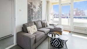 A modern living room with a grey sofa, a round black and white patterned coffee table, and a large window offering a city view. - Apartment Near North Sea and Lighthouse (Egmond aan Zee)
