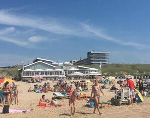 Un hôtel situé en bord de mer, reconnaissable à son grand panneau « Nautilus », doté d’une terrasse et d’une plage de sable où l’on peut voir des gens et des chaises longues.