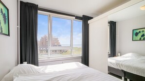 A hotel room with two beds, a large window with curtains, and a view of buildings and a beach. - Apartment Near North Sea and Lighthouse (Egmond aan Zee)