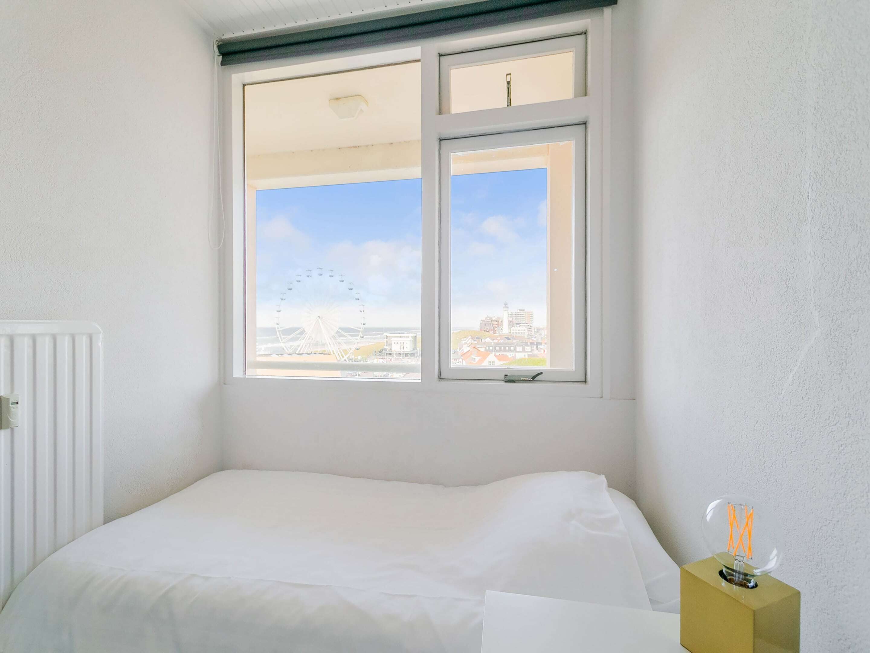 Ein Schlafzimmer mit Bett, Heizkörper und Fenster mit Blick auf eine Stadtlandschaft und ein Riesenrad.