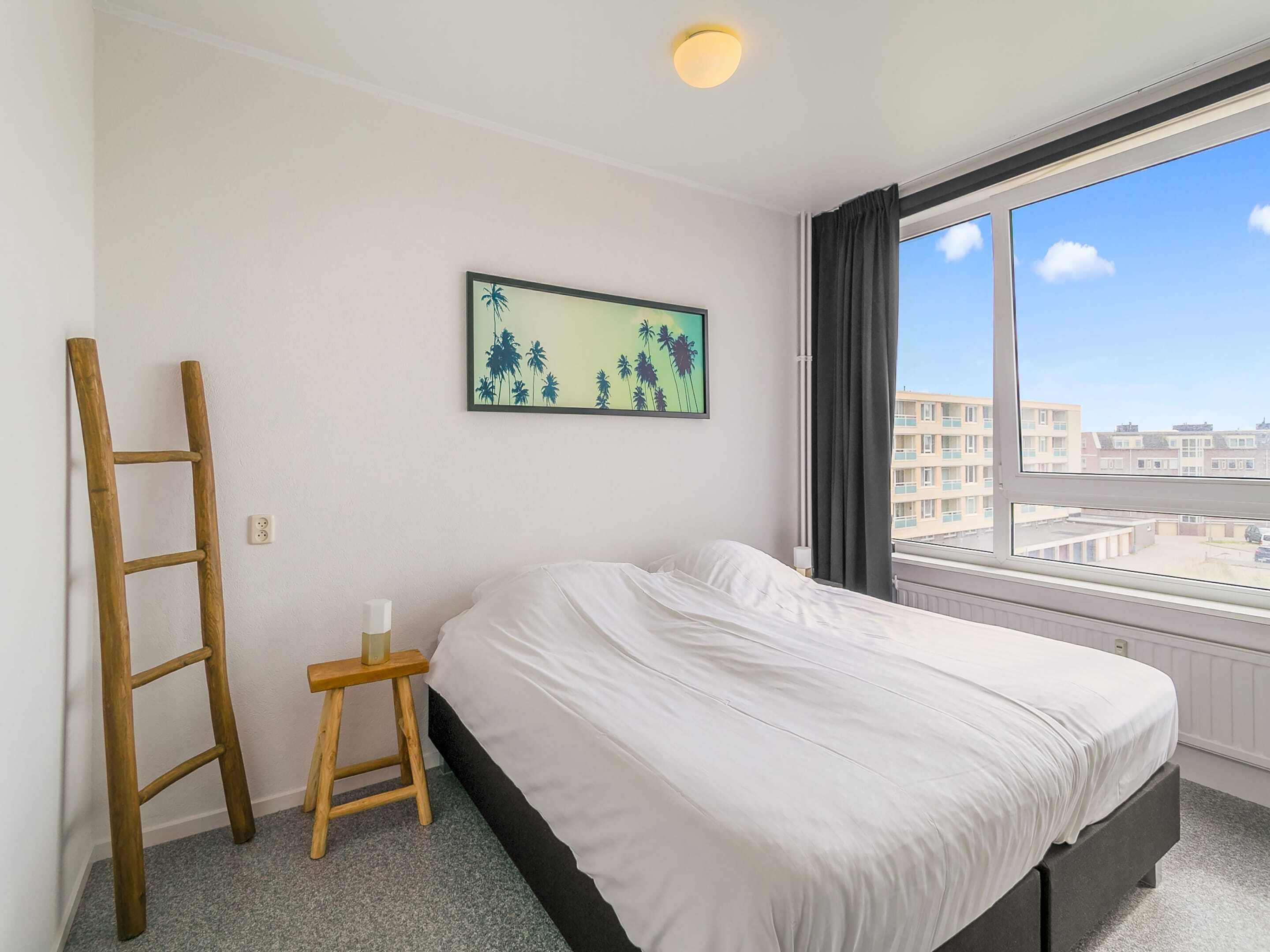 A bedroom with a bed, a wooden ladder, a small stool, a painting of palm trees, and a window with a city view.