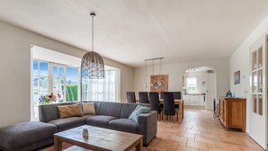 A spacious living area with a large sectional sofa, a wooden coffee table, and a dining table with chairs. There is a kitchen in the background with white cabinets and a view of the outdoors through large windows.