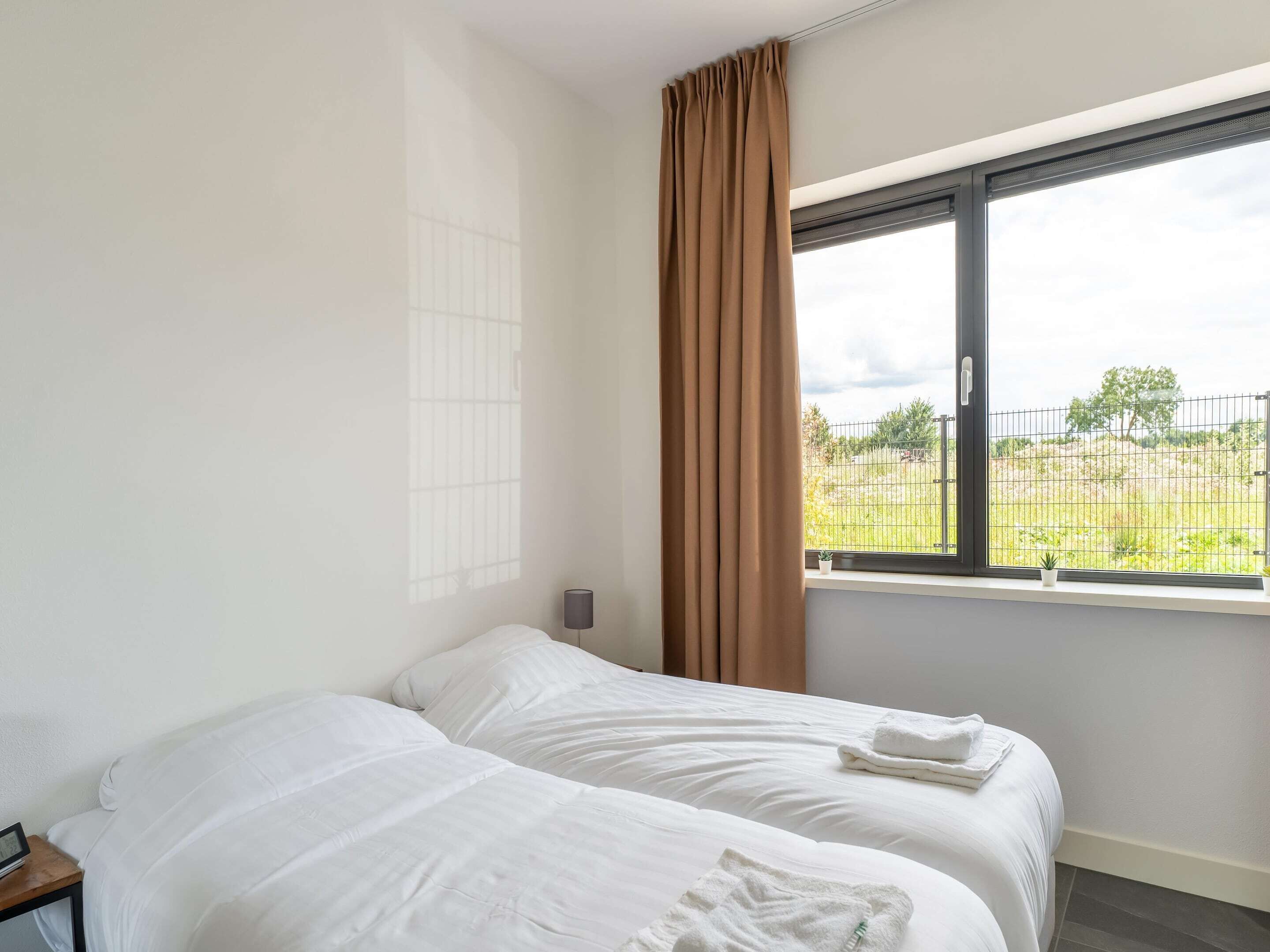 Habitación de hotel con dos camas, una ventana que da a un área exterior cercada y una mesita de noche con una lámpara.