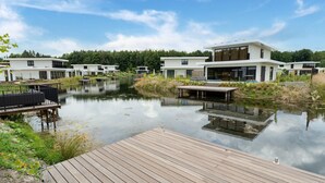 A modern residential area with a wooden dock extending into a calm water body, surrounded by lush greenery and contemporary white houses. - Villa in Zeewolde Near Flevostrand Beach (Zeewolde)