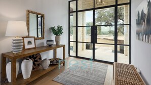 A modern interior with a wooden console table, a floor lamp, a mirror, and a large glass door leading to an outdoor area. - ***NEW*** INFINITY POOL | HOT TUB | Views | Pool Table | Fire Pit | 16 Queen Beds (Austin)