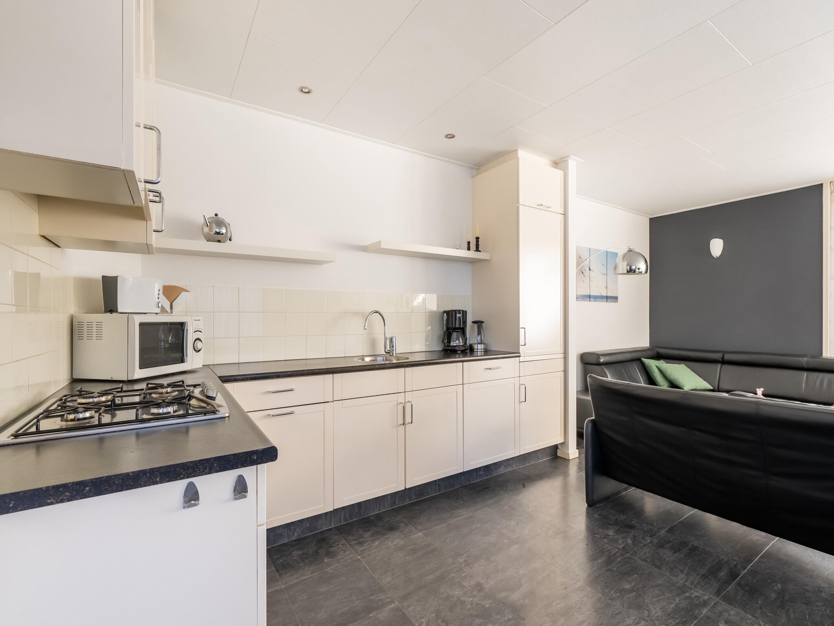 A modern kitchen with a gas stove, microwave, and coffee maker. There is a black couch and a dark wall in the adjacent living area.