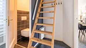 Une échelle en bois dans un couloir menant à une salle de bain carrelée de carreaux foncés et dotée de toilettes blanches.