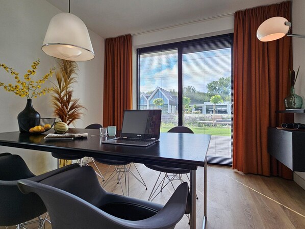 A modern dining area with a laptop on a table, black chairs, a vase with yellow flowers, and a view of a house through large windows.