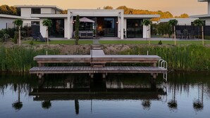 Una casa moderna con un muelle que se adentra en un lago tranquilo, rodeada de exuberante vegetación y un cielo despejado.