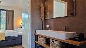 Una habitación de hotel moderna con una cama grande, una tocador de madera con un lavabo blanco y un espejo amplio. Hay cestas de mimbre debajo del tocador y una ventana con cortinas.