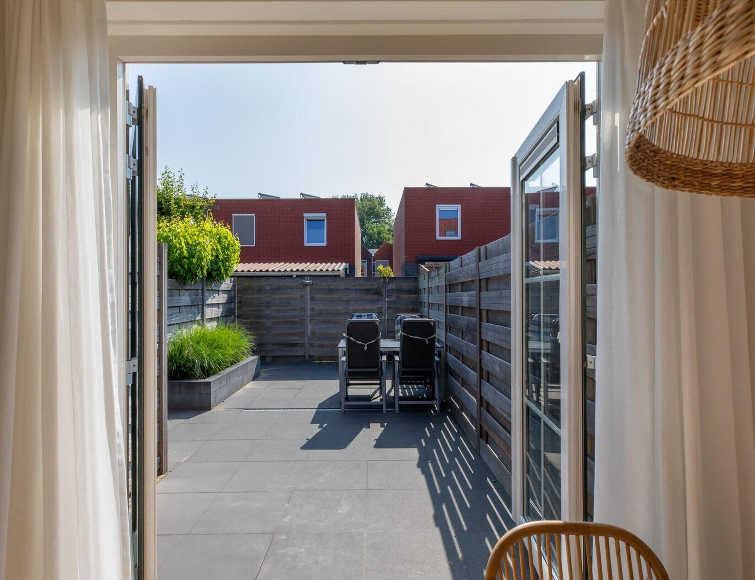 Un espace extérieur aménagé en terrasse, avec une table et des chaises, un fauteuil en osier et, en arrière-plan, une vue sur un bâtiment en briques rouges.