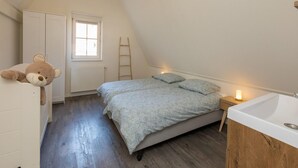 Quarto com teto inclinado, cama com manta estampada, abajur, escada de madeira, urso de pelúcia e janela com persianas.