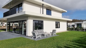 Una casa moderna de dos pisos con techo plano, ventanas amplias y un patio con mobiliario exterior.