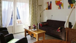 Un salon comprenant un canapé, une table basse en bois et une table à manger. Les murs sont ornés de tableaux abstraits et il y a une fenêtre avec des rideaux.