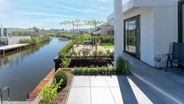 A modern outdoor patio with a view of a canal, wooden deck, and a garden area with plants and a small table.