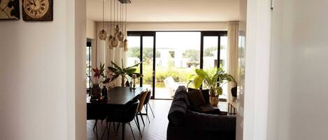 A modern living room with a dark sofa, a dining table with chairs, and a large window offering a view of greenery outside.