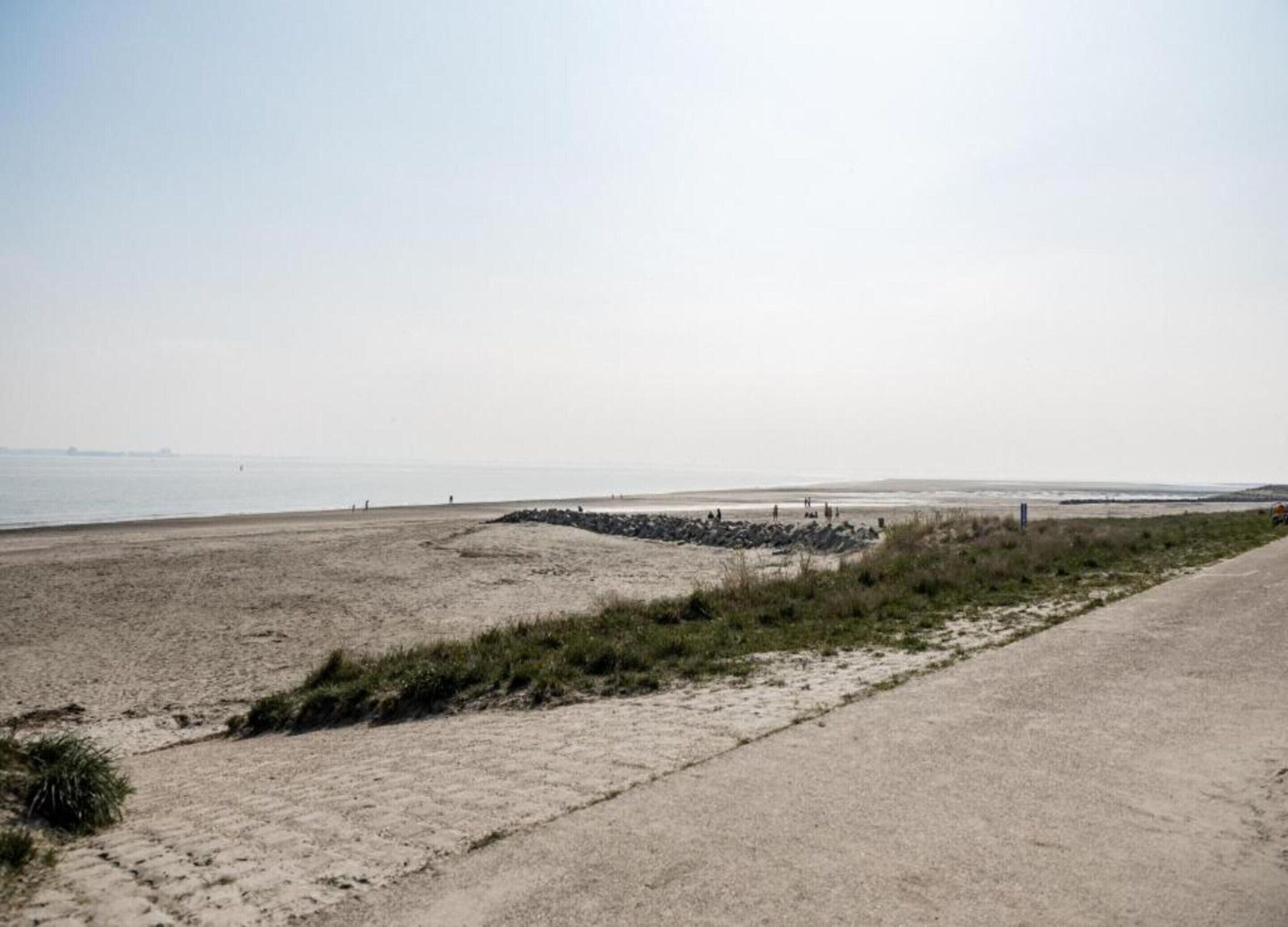 Uma praia com um caminho de concreto que leva à água, uma área rochosa e vista distante do mar.